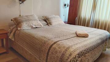 A neatly made bed with patterned bedding and two pillows, a bedside table with a lamp, and a window with curtains.
