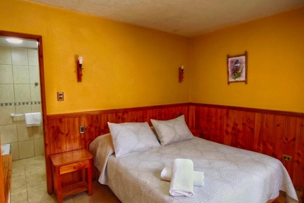 Een slaapkamer met een houten hoofdbord, een bed met een witte spreideken, twee kussens, een nachtkastje van hout en een ingelijste foto aan de muur.