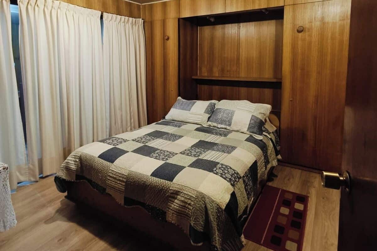 Een slaapkamer met houten lambrisering, een bed met ruitdessin en een raam met gordijnen.