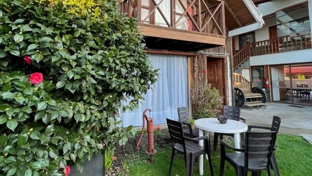 A wooden building with a balcony, a round table with chairs, and a well-maintained garden with a large bush and a flower pot. - Hostal & Cabañas Don Juan (Villarrica)