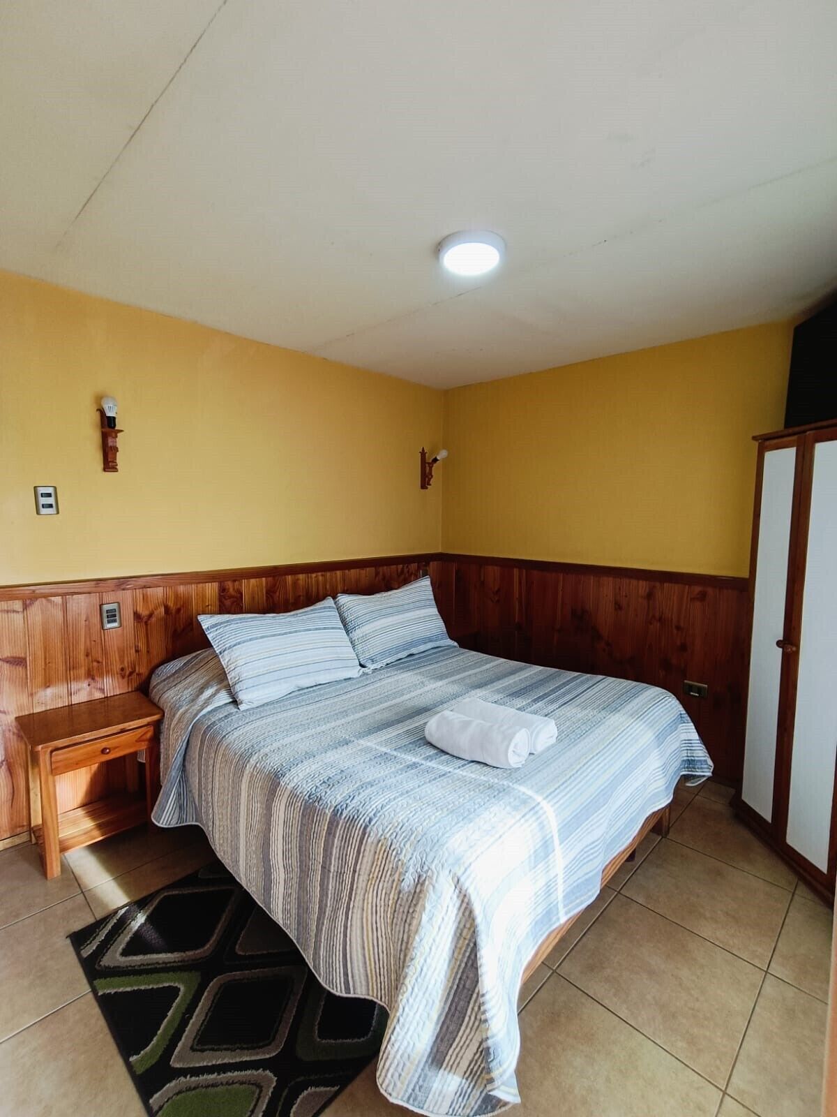 A single-bed room with wooden paneling, a bedside table, a nightstand, a patterned rug, and a wardrobe.