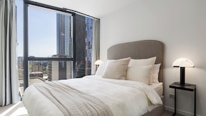 Une chambre d’hôtel moderne avec un grand lit, une table de chevet avec une lampe et une vue sur de hauts immeubles par une grande fenêtre.