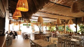 An indoor restaurant with thatched ceiling, wicker pendant lights, and potted plants. Tables and chairs are arranged for dining. - Maria Apartments by Checkin (Hersonissos)