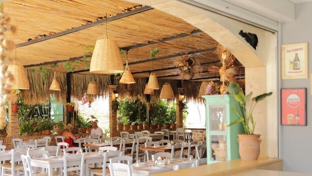 An outdoor restaurant with white tables and chairs, hanging wicker lamps, and potted plants. - Maria Apartments by Checkin (Hersonissos)