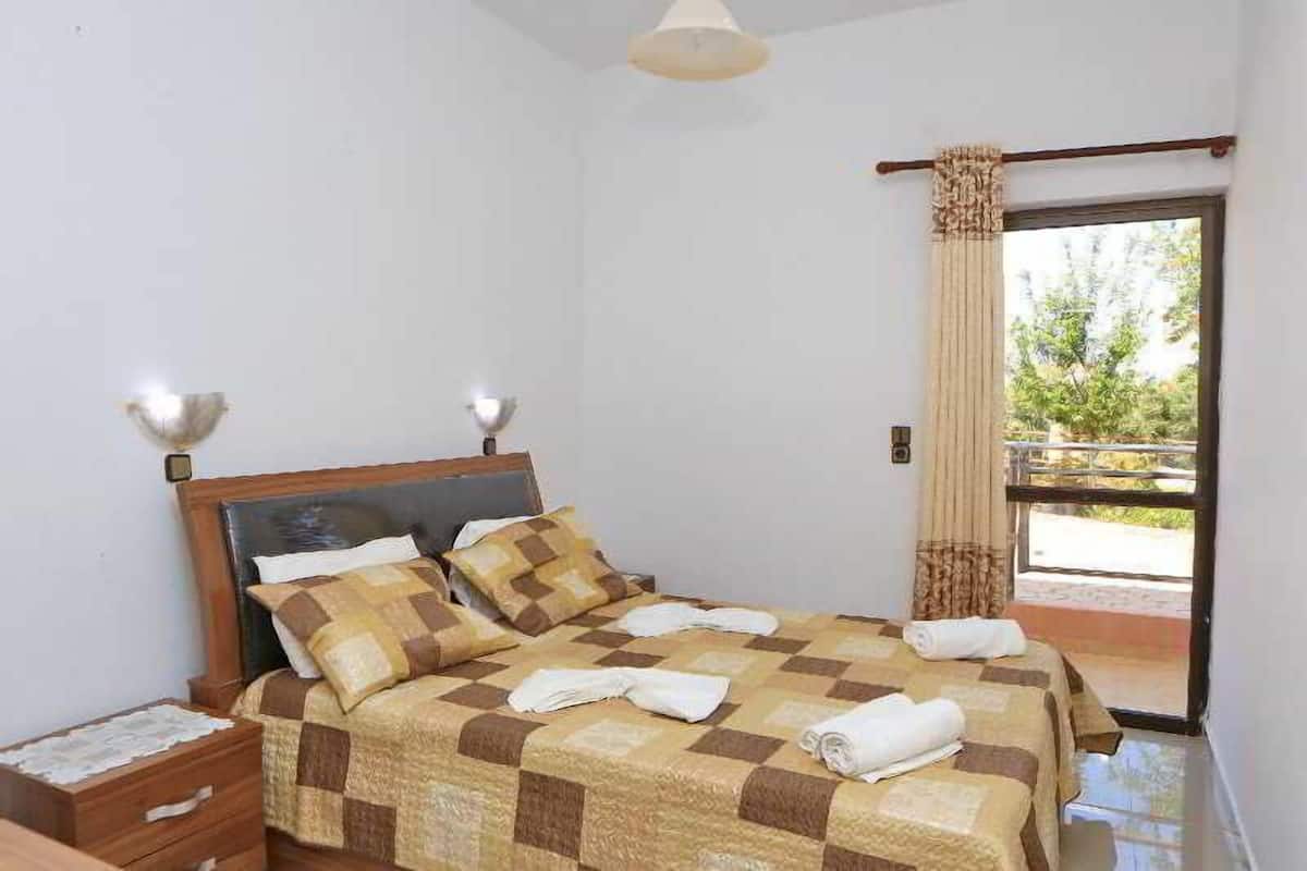 Un dormitorio con cama, mesitas de noche, ventana con cortinas y vistas al exterior.