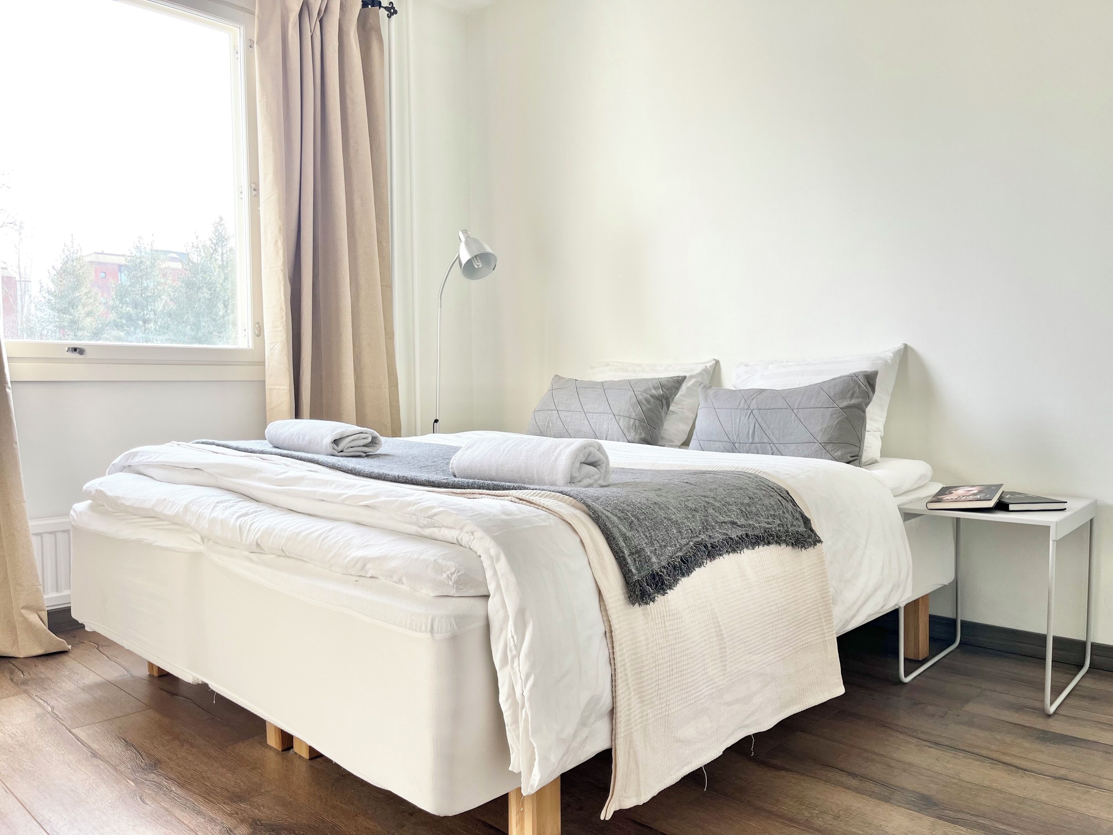 A neatly made bed with white linens and a gray throw blanket, a bedside table with a book, and a window with curtains.