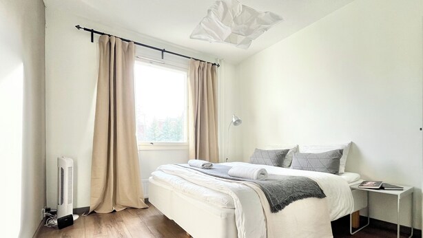 A modern bedroom with a large bed, a bedside table, a window with curtains, and a ceiling light fixture. - City Home Finland (Tampere)