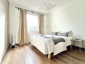 A modern bedroom with a large bed, a bedside table, a window with curtains, and a ceiling light fixture. - City Home Finland (Tampere)