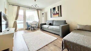 A modern living room with a grey sofa, a white desk, and a television. There is a dining table with chairs and a window with curtains. A painting is on the wall. - City Home Finland (Tampere)