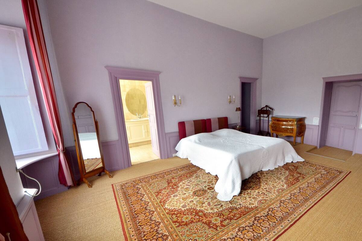 A bedroom with a large bed, a dresser, a mirror, and a door leading to another room.