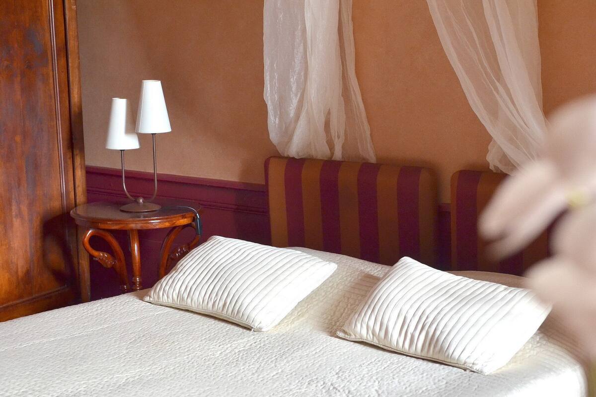 A neatly made bed with white linens and pillows, a small round wooden side table with a lamp, and a striped upholstered chair.