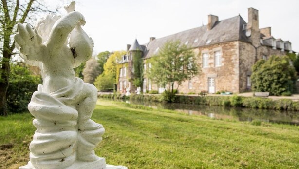 A white sculpture of a figure with a twisted body, set on a pedestal, in front of a historic stone building with multiple chimneys and a pond. - Château de la Motte Beaumanoir (Pleugueneuc)
