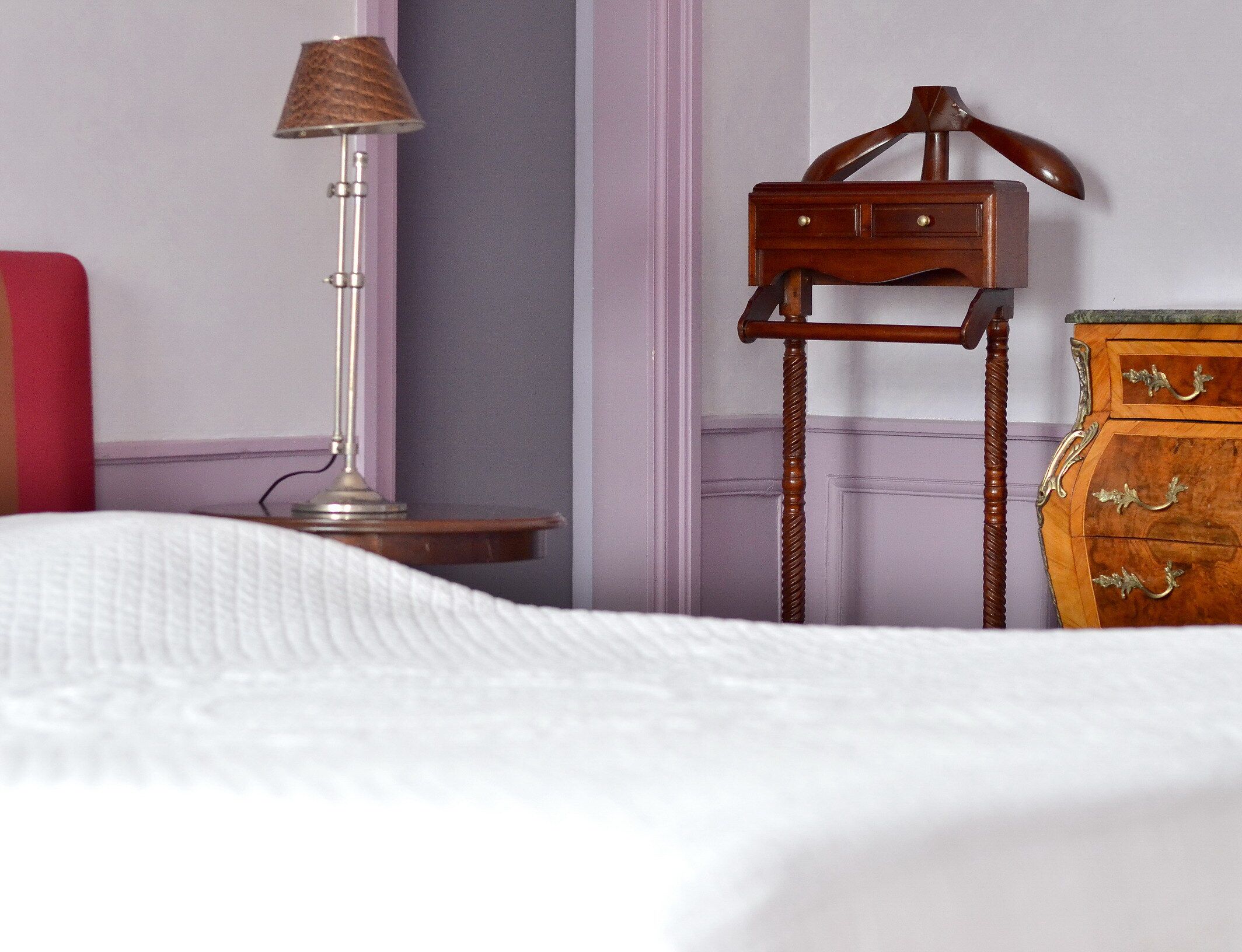 Un letto rifatto con un copriletto bianco, un piccolo comodino in legno con una lampada e una cassettiera in legno con cassetti.