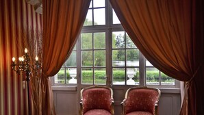 A room with two chairs facing a window with curtains, a chandelier, and a view of a garden and water feature. - Château de la Motte Beaumanoir (Pleugueneuc)