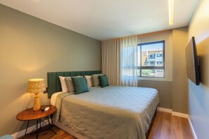 Habitación de hotel con cama, mesita de noche con lámpara, ventana con cortinas y televisor montado en la pared.