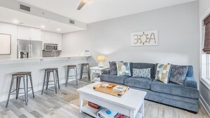 A modern living space with a kitchen, bar stools, a sofa with cushions, and a coffee table. - Cassine Way 303 - Seagrove + Sunsets (Santa Rosa Beach)