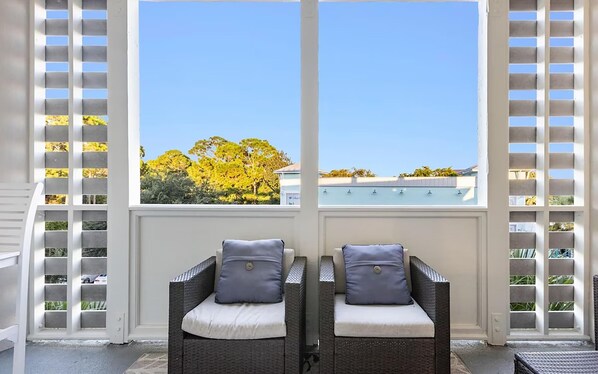 Une pièce avec deux fauteuils en osier dotés de coussins, une vue sur des arbres et un bâtiment, le tout visible à travers de grandes fenêtres à cadres blancs.