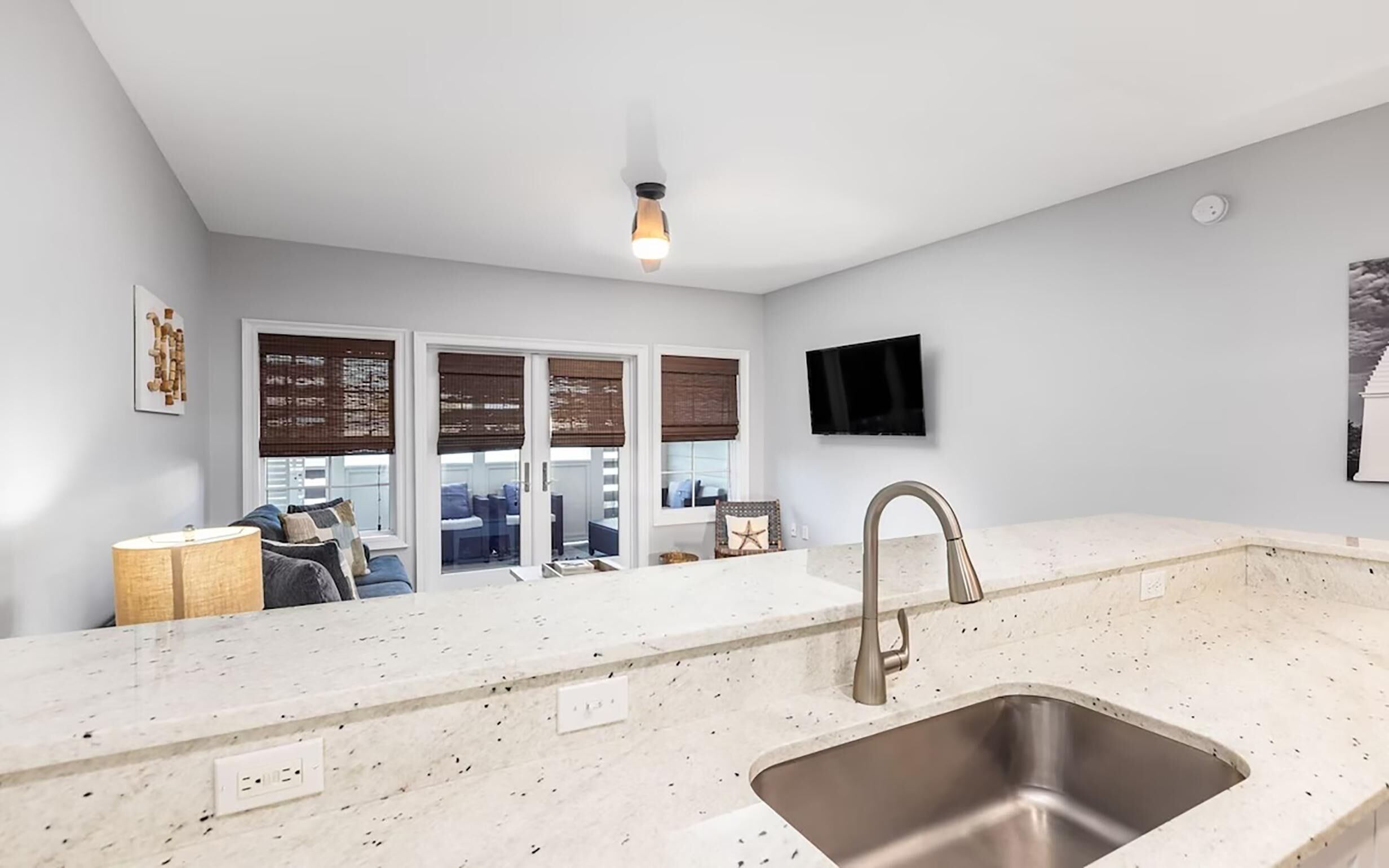 A modern kitchen with a stainless steel sink, a flat-screen TV mounted on the wall, and a large window with blinds.