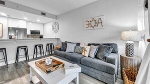 A modern living room with a grey sofa, a wooden coffee table, and a kitchen area with white cabinets and a stainless steel refrigerator. - Cassine Way 303 - Seagrove + Sunsets (Santa Rosa Beach)