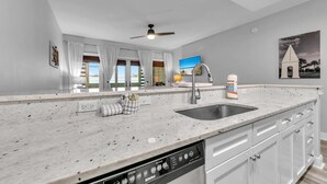 A kitchen with a large island, a built-in dishwasher, and a view of the outdoors through large windows. - Cassine Way 303 - Seagrove + Sunsets (Santa Rosa Beach)