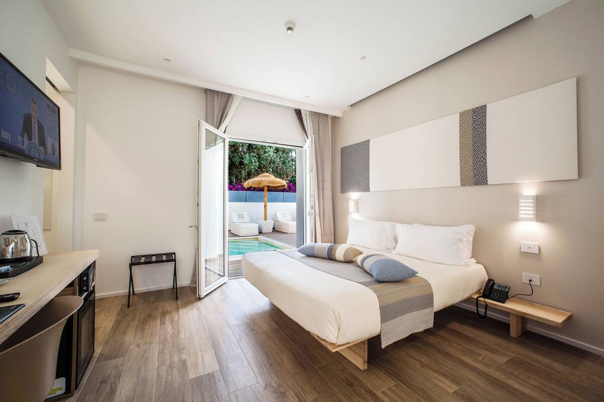A modern hotel room with a large bed, a desk with a kettle, a small table, and a view of a swimming pool.