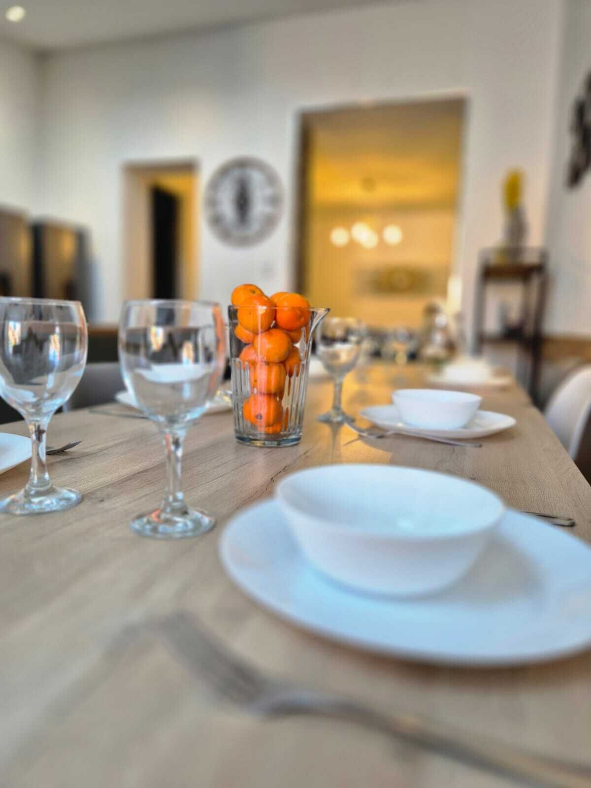 Una mesa de comedor dispuesta con platos blancos, vasos transparentes y una jarra de vidrio con naranjas.