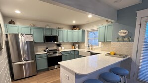 A modern kitchen with teal cabinets, stainless steel appliances, a marble island with bar stools, and a window with blinds.