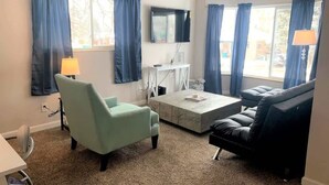 Uma sala de estar com uma poltrona verde clara, um sofá de couro escuro, uma mesa de centro e uma TV fixada na parede.