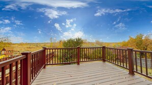 A wooden deck with a brown railing, overlooking a natural landscape with trees and a clear blue sky.
