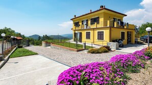 Un edificio amarillo de dos pisos con un balcón, un camino de entrada pavimentado y un jardín de flores.
