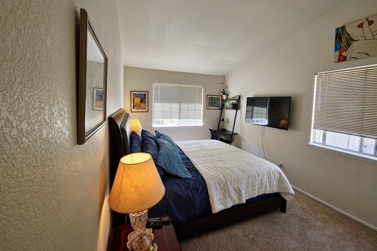 A bedroom with a bed, a bedside table with a lamp, a TV mounted on the wall, and a window with blinds.