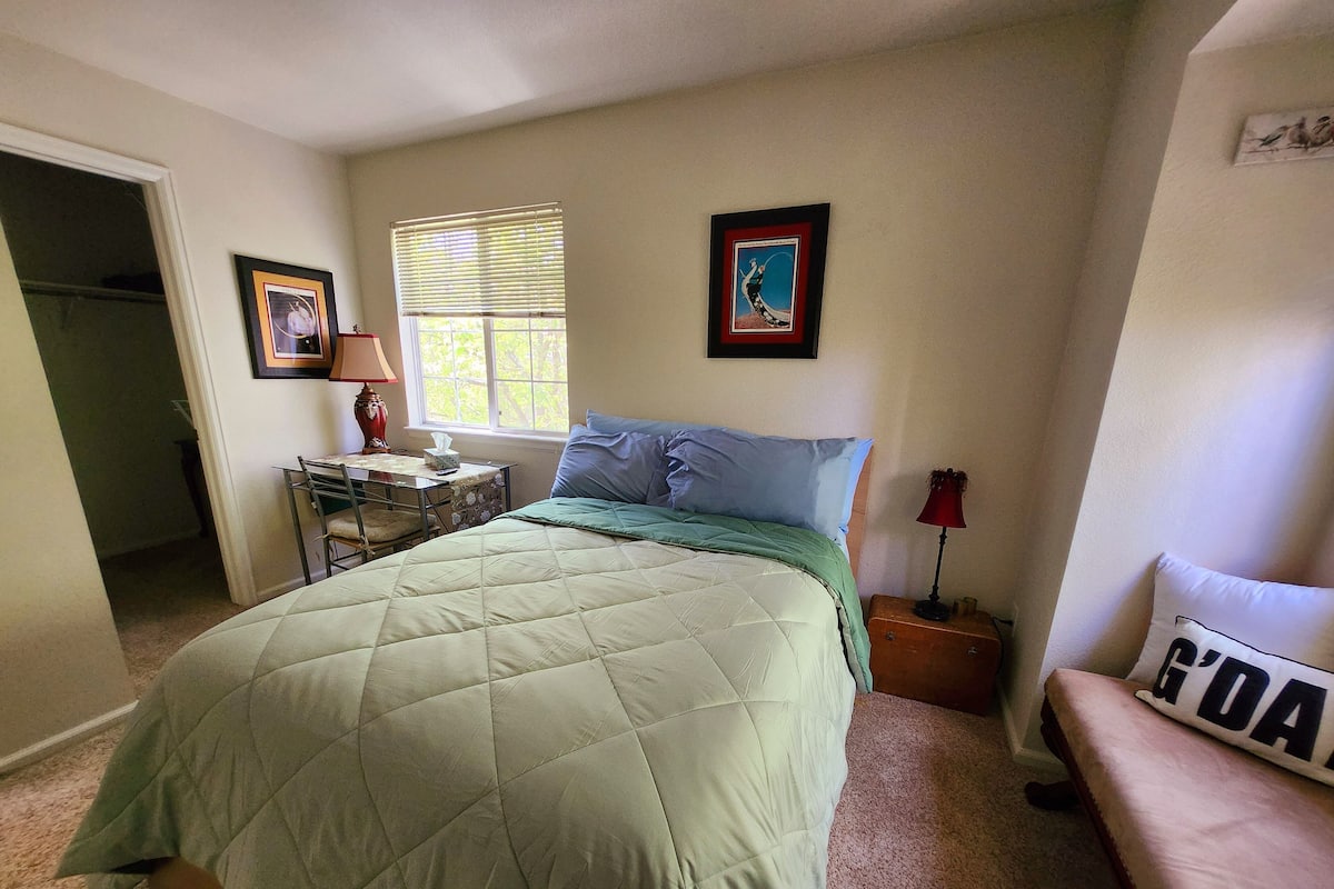 A bedroom with a bed, a desk, a chair, a window with blinds, and a door.