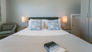 A neatly made bed with a book titled 'Breath' by James Nestor, two pillows with sailboat designs, a grey upholstered chair, and two bedside lamps. - Pen Tywyn - sleeps 7 guests  in 3 bedrooms (Rhosneigr)