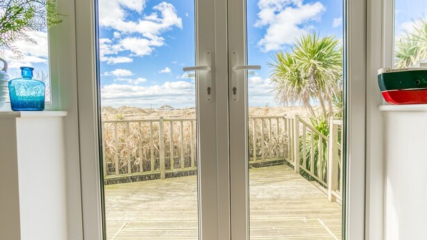 A view through a glass door to a wooden deck with a railing, overlooking a sandy landscape and palm trees. - Pen Tywyn - sleeps 7 guests  in 3 bedrooms (Rhosneigr)