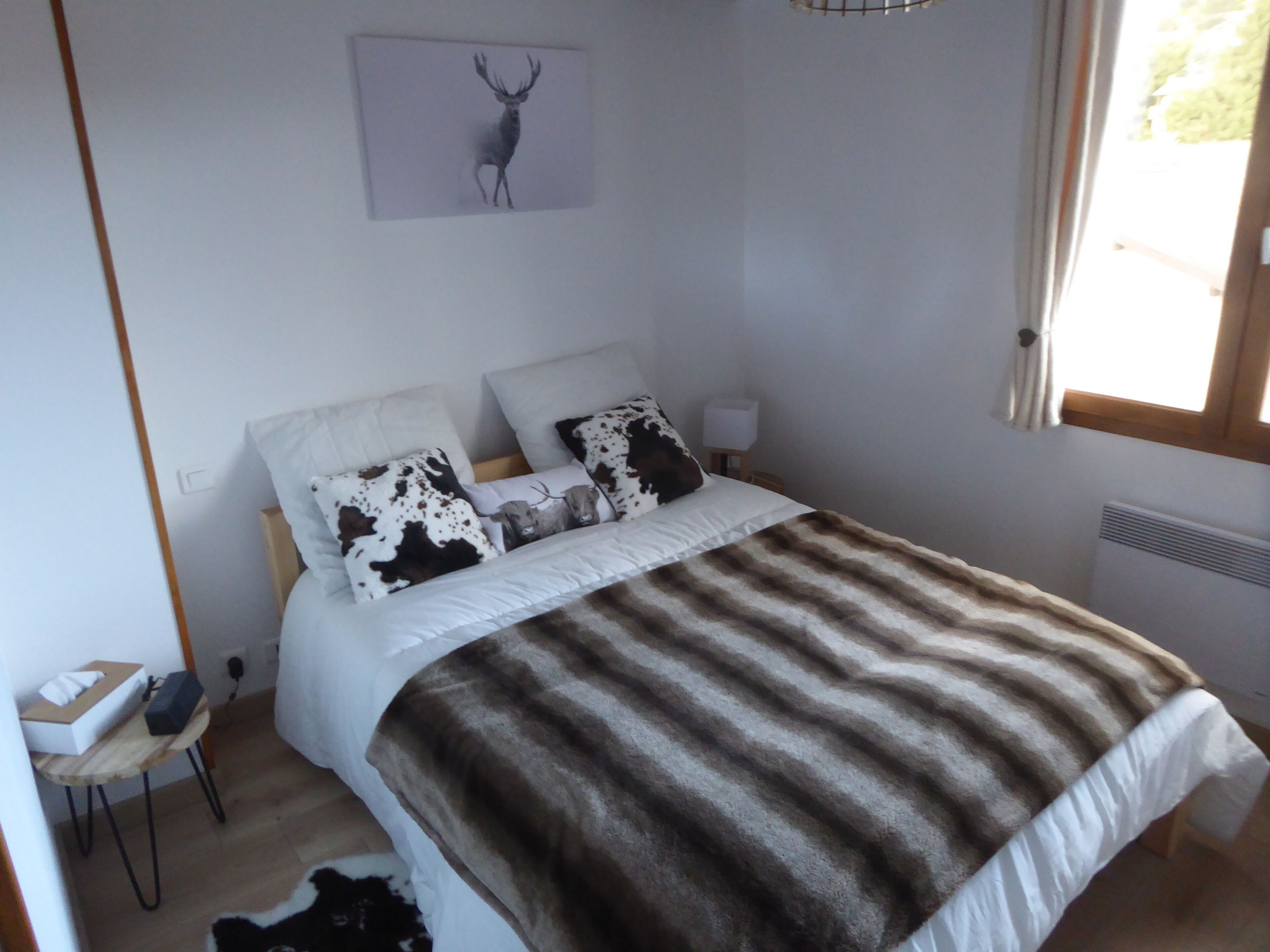 A bedroom with a bed, white bedding, two decorative pillows, a blanket, a bedside table with a lamp, a small rug, and a wall-mounted picture of a deer.