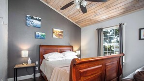 A bedroom with a wooden bed, a nightstand with a lamp, and a window with curtains. - Tiled Shower, Walk to Main, Kitchenette, Smart TV (Fredericksburg)
