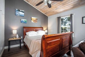 A bedroom with a wooden bed, a nightstand with a lamp, and a window with curtains. - Walk to Main, Tiled Shower, Smart TV, Queen Bed (Fredericksburg)