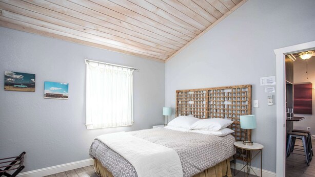 A bedroom with a wooden ceiling, a bed with a headboard, a window with curtains, and a small table with a lamp.