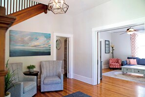 A spacious living room with hardwood floors, a grey armchair, a blue sofa, and a painting of a sunset. - Chesapeake Charm Estate, Two Blocks from the Beach (Cape Charles)