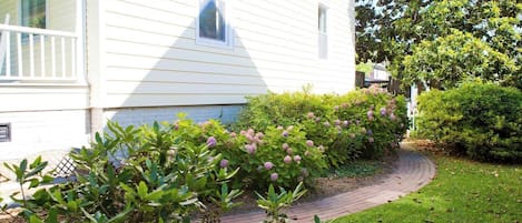 Een wit huis met een met baksteen bestrate looppad die leidt naar een verzorgde tuin met hortensiakweest.'