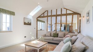 A spacious living room with a high ceiling, exposed wooden beams, a large sofa, a coffee table, and a view into the kitchen. - The Hide (Colchester)