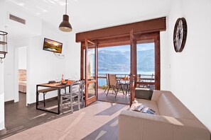 A modern living room with a sofa, dining table, and chairs, a television mounted on the wall, and a large window overlooking a scenic view.