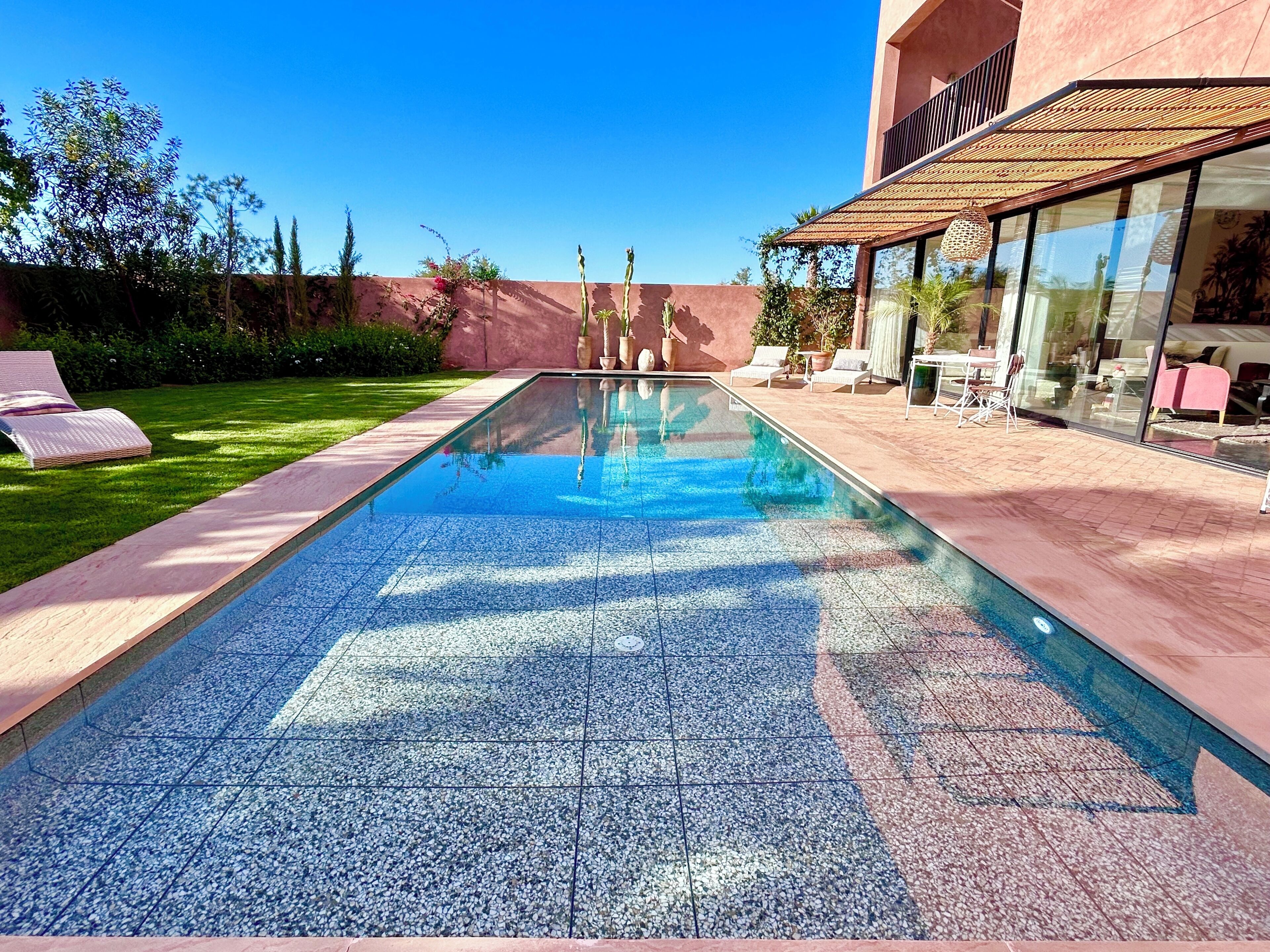 A rectangular swimming pool with a pebble pattern, surrounded by a patio area with lounge chairs and a glass-enclosed section with furniture.