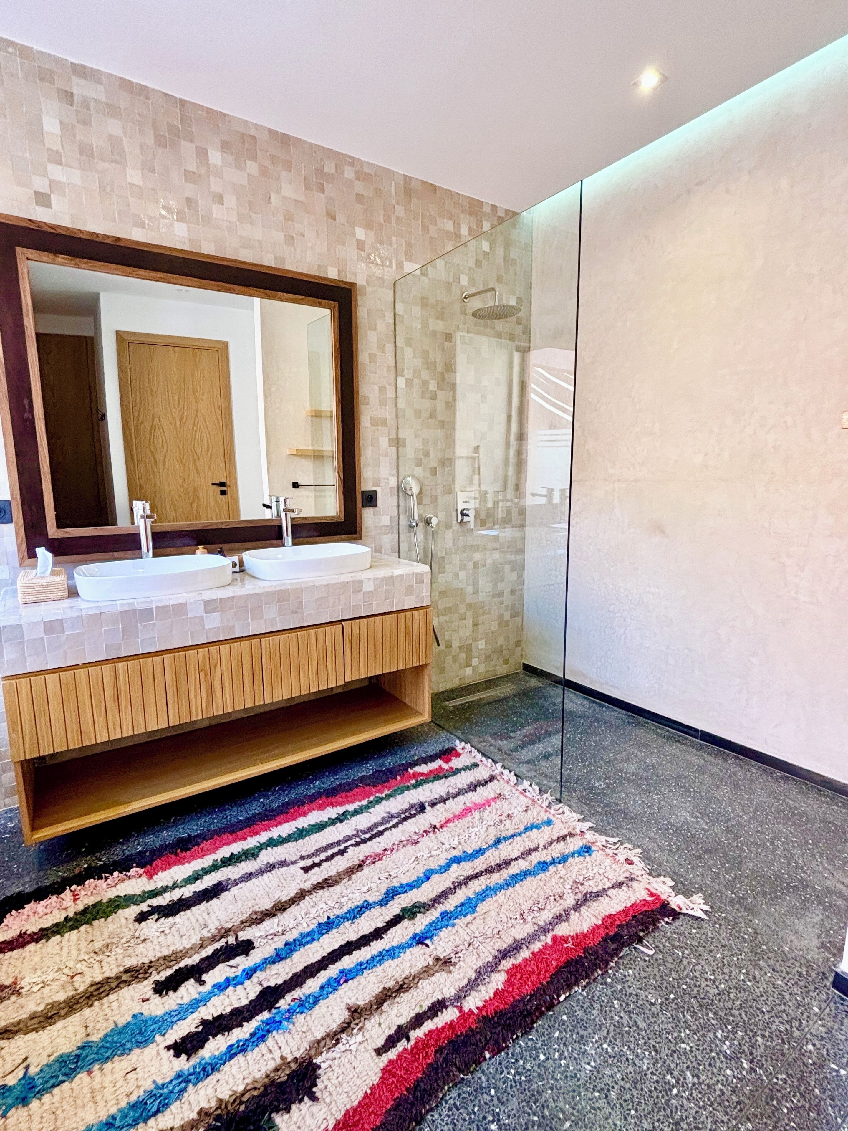 Une salle de bain moderne avec un grand miroir, deux lavabos, une douche et un tapis rayé aux couleurs vives.