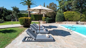 A poolside area with lounge chairs and umbrellas, surrounded by well-manicured bushes and trees.