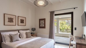 A modern bedroom with a large bed, a desk, and a TV. There are framed artworks on the wall and a view of the outdoors through a sliding door.