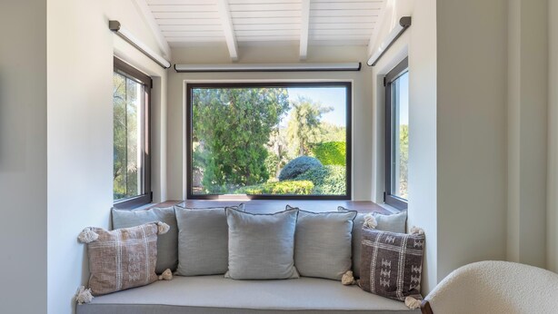 A cozy seating area with a bench and cushions, large windows offering a view of greenery outside.