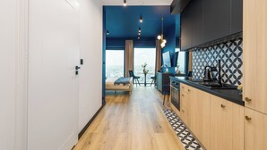 A modern hotel room with a wooden floor, a bed, a dining area with a table and chairs, a kitchen with a black countertop, and a large window offering a city view. - Apartament Bulvar - by Kairos Apartments (Katowice)