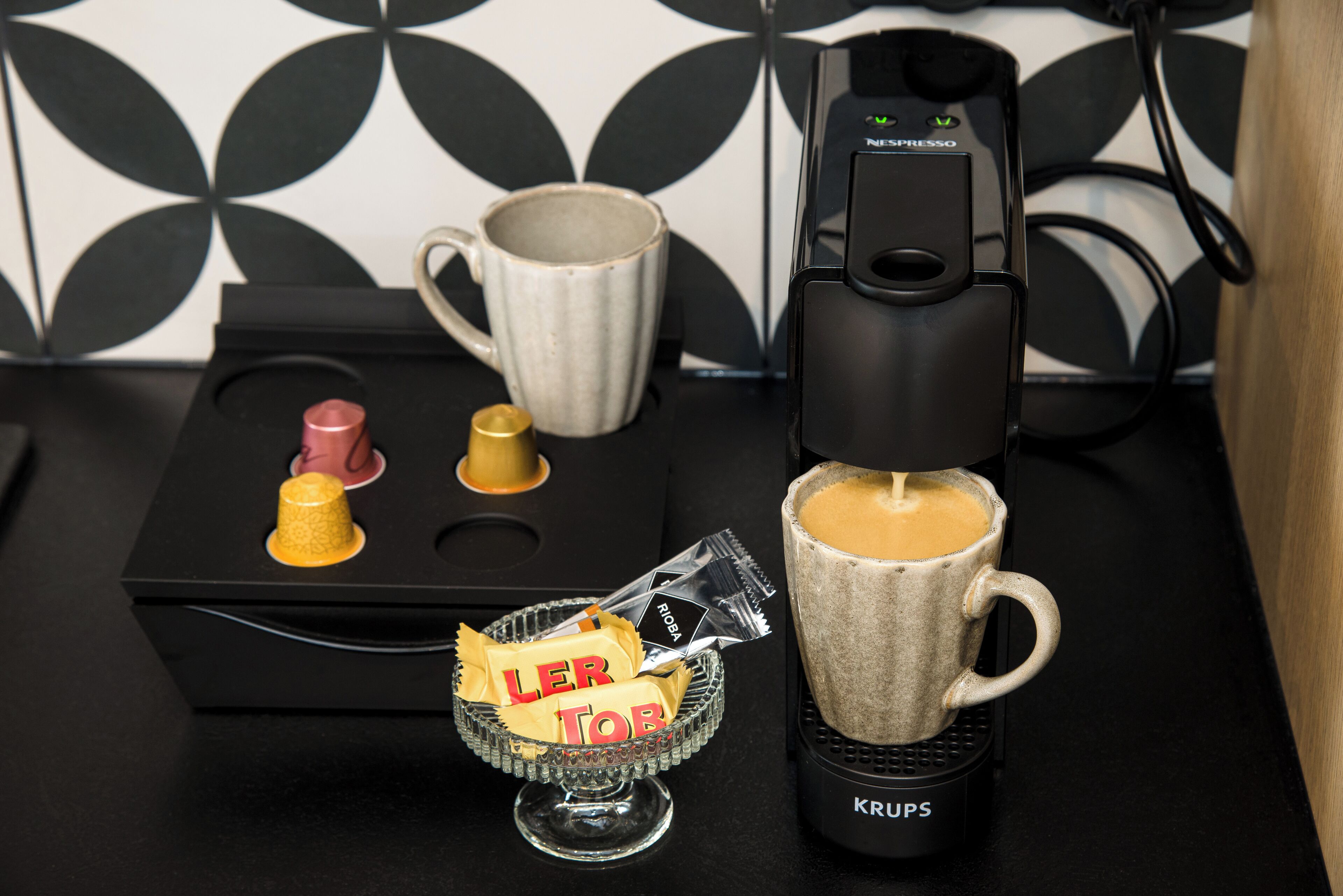 Une machine à café, une tasse, un verre et une petite assiette contenant des dosettes de café et un sachet étiqueté « LER TOR » sur un plan de travail noir.
