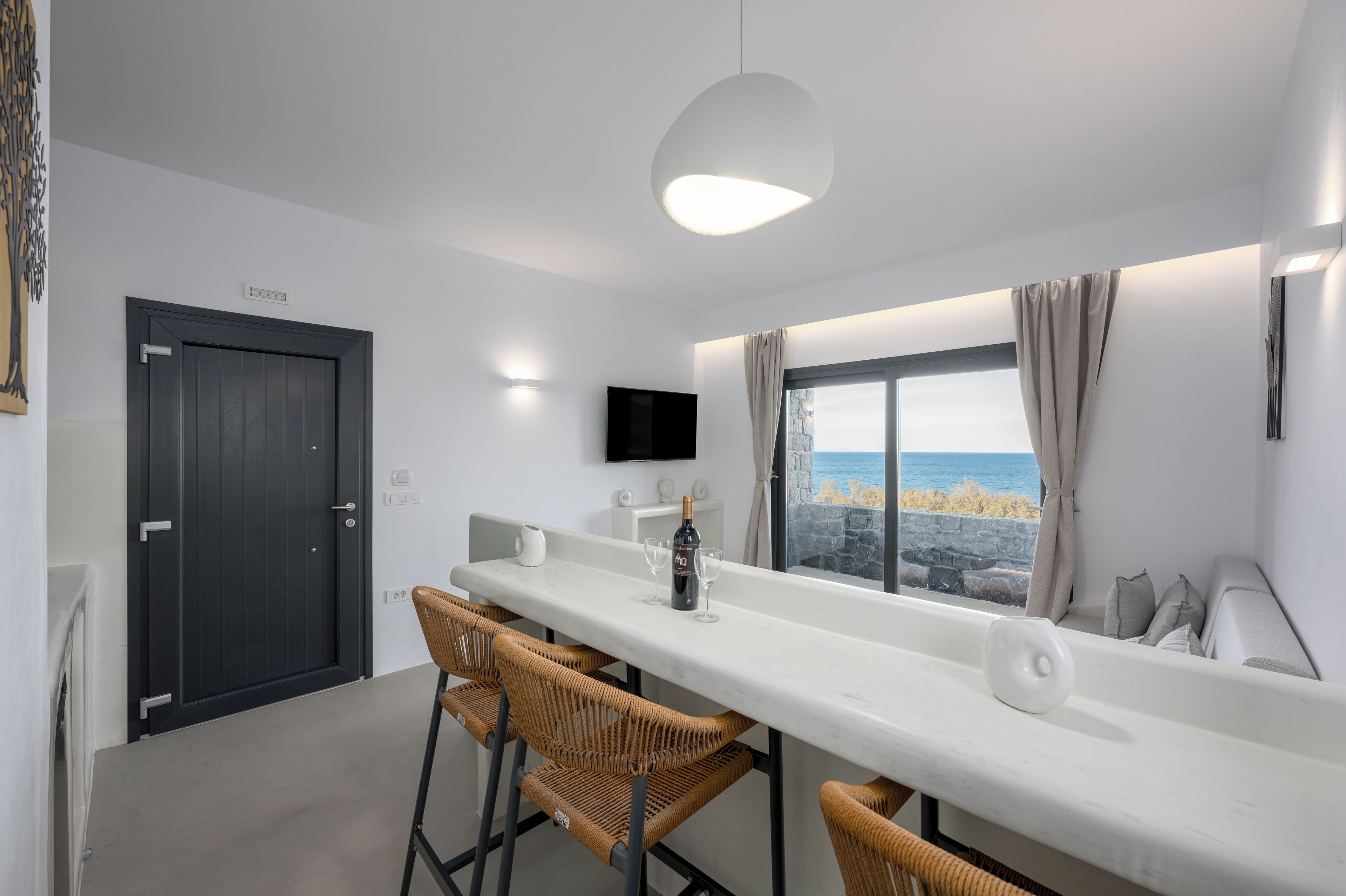 A modern kitchen with a long white countertop, wicker chairs, a dark door, and a window with a view of the sea.